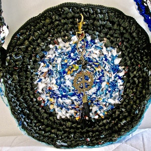Pu&ograve; includere: Una borsa rotonda in crochet nero con una tracolla in crochet blu e bianca. La borsa ha un ciondolo a forma di segno della pace.