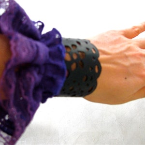K&ouml;nnte beinhalten: Ein lila-wei&szlig;es, gekr&auml;useltes Stoffarmband mit einer schwarzen Spitzenmanschette. Das Armband wird am Handgelenk getragen.