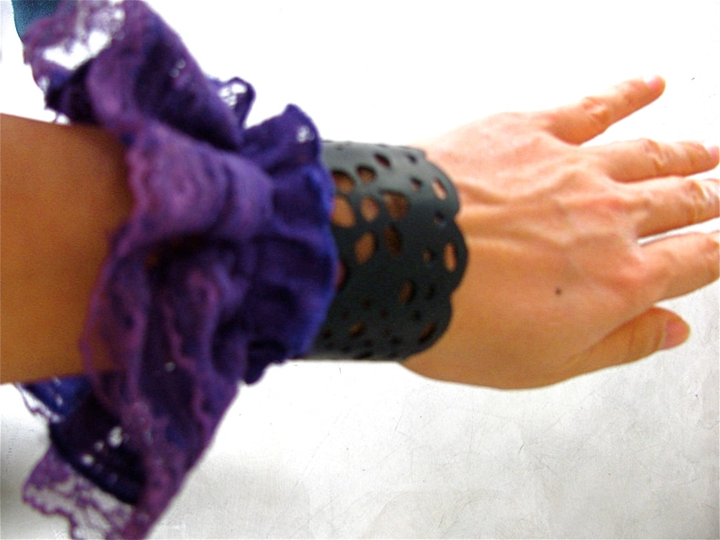 K&ouml;nnte beinhalten: Ein lila-wei&szlig;es, gekr&auml;useltes Stoffarmband mit einer schwarzen Spitzenmanschette. Das Armband wird am Handgelenk getragen.