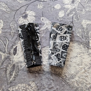 May include: Two black and white patterned arm cuffs. One cuff is laced with a black ribbon. The other cuff features a repeating pattern of skulls, moons, stars, and other occult symbols. The cuffs are displayed on a gray floral patterned surface.
