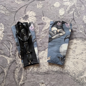 May include: Two blue fabric arm cuffs with black ribbon and silver-tone metal rings. One cuff features a black ribbon design, while the other displays a gothic illustration of a woman, bats, and a full moon.