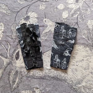 May include: Two black fabric arm cuffs with white Halloween-themed illustrations. One cuff has black straps and buckles. The other cuff features a skull, spiderwebs, and potion bottles. The word "Halloween" is also visible.