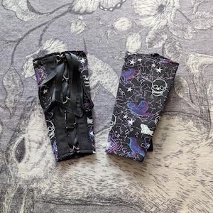 May include: Two black arm cuffs with a celestial and skull print. One cuff has a black ribbon lace-up design. The other cuff features a print of birds, stars, and constellations. The cuffs are made of a dark fabric with purple and blue accents.
