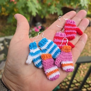 May include: A selection of handmade crochet mitten earrings in various colours, including blue, white, pink, red, and orange. Each earring features a miniature mitten design with silver-coloured ear wires. The mittens have striped and solid colour patterns.