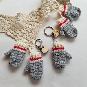 May include: Four grey crocheted mitten keychains with red and white trim. Each keychain has a silver key ring and a small wooden heart charm. One charm has the text "Always & Forever" engraved on it.