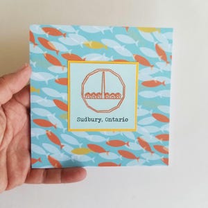 May include: A blue card with a yellow border and a white circle with a stylised image of a building and the text "Sudbury, Ontario". The background is a pattern of orange and yellow fish on a blue background.