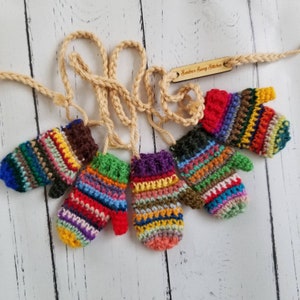 May include: A garland of five miniature crocheted mittens in various multicoloured striped patterns, each attached to a single beige crocheted string. A small wooden tag with the text Sandra's Sassy Stitches is attached to the string.
