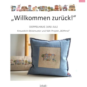 May include: A blue cushion featuring a stitched house design, part of a cross-stitch and sewing project. The text "Willkommen zurück!" is at the top, with "DOPPELHAUS JUNI-JULI" below. The cushion has a light-coloured centre with a house and tree design.