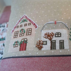 Könnte beinhalten: Ein dekoratives Kissen mit Kreuzstichdesign, das zwei Häuser mit roten Dächern und grünen Kränzen zeigt. Das Kissen hat einen beigen Hintergrund mit weißen Punkten und einen rot-weiß gestreiften Boden.
