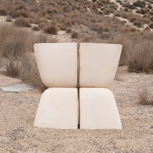 May include: A white concrete sculpture of a chair with a simple, modern design. The chair is sitting on a sandy beach with dry grass in the background.