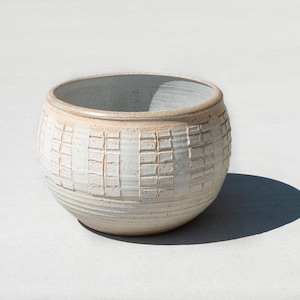 May include: A handcrafted, off-white ceramic bowl with a textured, grid-like pattern. The bowl has a rounded shape with a slightly flared rim and a dark interior. The bowl is sitting on a light-colored surface.