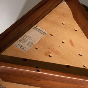 May include: Close-up of a wooden triangular table. The table has a dark brown frame and a light brown surface with several holes. A label is affixed to the surface with text that reads "DO NOT REMOVE THIS TAG".