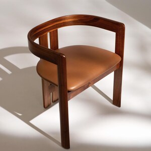 May include: A wooden armchair with a curved backrest and armrests. The seat is upholstered in brown leather. The chair has a minimalist design with clean lines and a warm, inviting aesthetic. The chair is made of wood and leather.