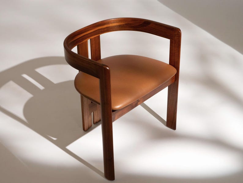 May include: A wooden armchair with a curved backrest and armrests. The seat is upholstered in brown leather. The chair has a minimalist design with clean lines and a warm, inviting aesthetic. The chair is made of wood and leather.