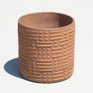 May include: A cylindrical, terracotta-colored ceramic pot with a textured, woven-like pattern. The pot has a wide opening and a rounded rim. The exterior features a repeating geometric design, adding visual interest to the piece. The pot is suitable for plants or decorative use.