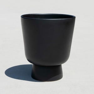 May include: A matte black ceramic planter with a rounded base and a wider top. The planter has a simple, modern design and is suitable for indoor or outdoor use. The planter is approximately 18 cm tall.