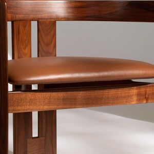 May include: A close-up of a wooden chair with a brown leather seat. The chair has a dark wood frame with a rectangular back and armrests. The seat is oval-shaped and appears to be made of leather. The chair is a piece of furniture.