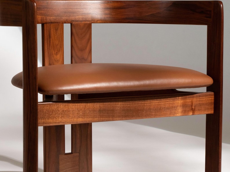 May include: A close-up of a wooden chair with a brown leather seat. The chair has a dark wood frame with a rectangular back and armrests. The seat is oval-shaped and appears to be made of leather. The chair is a piece of furniture.