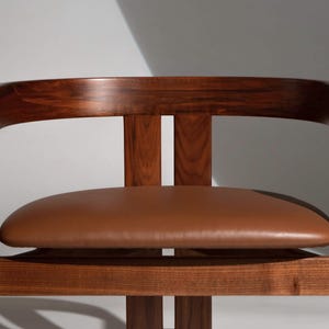 May include: A mid-century modern chair with a curved, dark wood frame and a brown leather seat. The chair has a minimalist design, showcasing the natural wood grain and the smooth texture of the leather. The backrest is curved for comfort.