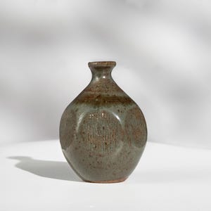 May include: A small, handcrafted ceramic vase with a rounded body and a narrow neck. The vase features a mottled glaze in shades of brown and green, with a textured surface. The vase is approximately 6 inches tall.