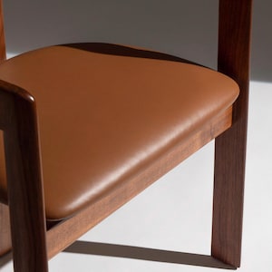 May include: Close-up of a wooden chair with a brown leather seat. The chair has a dark wood frame and a square seat. The seat is a warm, medium brown color. The chair is a modern design.