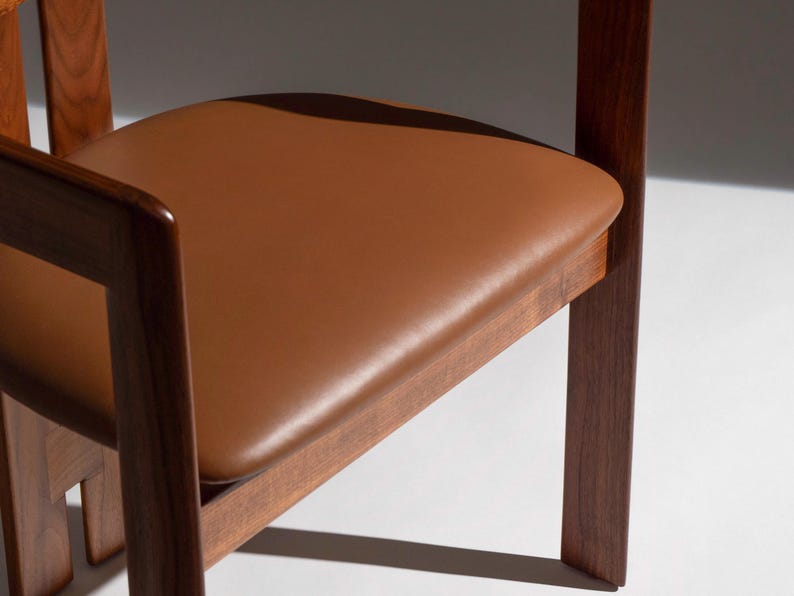 May include: Close-up of a wooden chair with a brown leather seat. The chair has a dark wood frame and a square seat. The seat is a warm, medium brown color. The chair is a modern design.