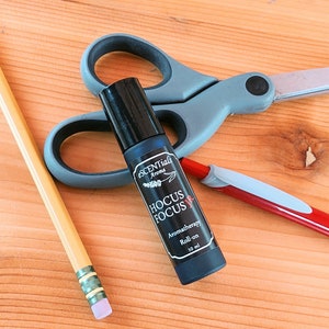 May include: A black roll-on bottle labeled "scentuals aroma hocus focus aromatherapy roll-on 10 ml" on a wooden surface with a pair of scissors and a red pen.