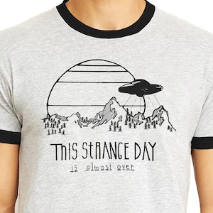 May include: A gray t-shirt with a black ringer. The shirt features a graphic of a UFO over a mountain range with the text "This Strange Day is almost over".