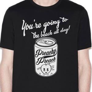 May include: Black t-shirt featuring a white graphic of a can of soda. The can reads "Peachy Peach" and depicts a cartoon peach with a face. The text "You're going to the beach all day!" is positioned above the can.