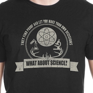 Könnte beinhalten: Schwarzes T-Shirt mit einem abgenutzten weißen Grafikdruck eines wissenschaftlichen Logos. Das Logo zeigt ein Atom, ein Becherglas, ein Mikroskop und den Text "They find proof and let you make your own decisions. What about science?"