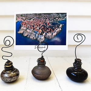 May include: Three decorative card holders featuring rounded bases in metallic, matte brown, and glossy black finishes. Each base is topped with a coiled wire stem designed to hold a rectangular postcard showing an aerial view of Dubrovnik, Croatia.