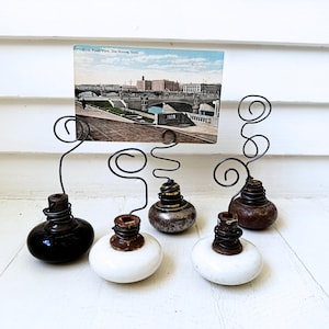 May include: A collection of vintage photo holders. Each holder features a decorative wire design with a bulb-shaped base in black, white, and metallic finishes. A postcard is held in one of the holders.