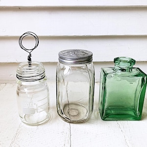May include: Three glass containers on a white surface. A small clear jar with a wire handle, a larger jar with a metal lid, and a square green glass bottle. The jars are empty, and the bottle has a unique shape.
