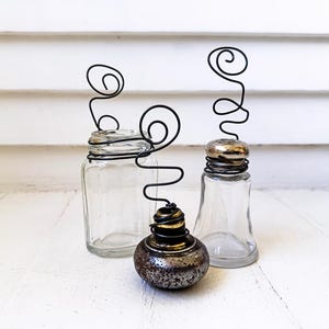 May include: Three decorative glass jars with black wire accents. The jars vary in shape and size, with one having a metal lid and another a decorative metal base. The wire forms a unique, swirling design above each jar.