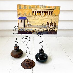 May include: A vintage metal photo holder with three bases, each supporting a wire frame. The bases are a rusty brown, a dark brown, and a black glass bottle. A postcard of Athens, Greece, is held in the frame. The text "Athens Sintagma" is visible.