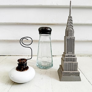 May include: A collection of decorative items including a white ceramic inkwell with a wire pen holder, a clear glass salt shaker with a black lid, and a silver metal replica of the Chrysler Building.