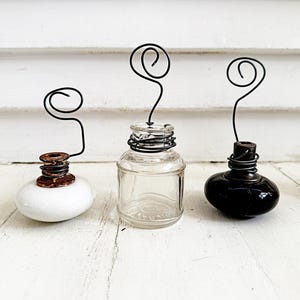 May include: Three vintage ink bottle holders. One is white, one is clear glass, and one is black. Each has a wire spiral design extending upwards. The holders are on a white wooden surface.