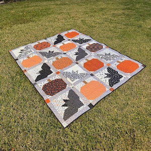 Könnte beinhalten: Ein Halloween-Quilt mit orangefarbenen Kürbissen und schwarzen Fledermäusen auf einem grau-weißen Patchwork-Hintergrund. Der Quilt besteht aus verschiedenen gemusterten Stoffen und hat einen schwarzen Rand.
