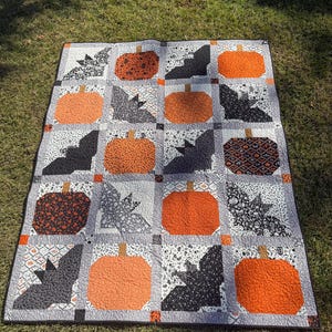 Könnte beinhalten: Ein Halloween-Quilt mit orangefarbenen Kürbissen und grauen Fledermäusen auf hellgrauem Hintergrund. Der Quilt besteht aus quadratischen Blöcken mit verschiedenen Mustern, darunter Blumen- und geometrische Designs. Der Quilt wird auf grünem Gras gezeigt.