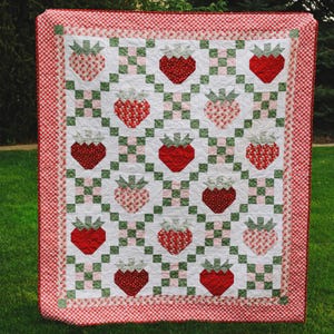 May include: A handmade quilt featuring a repeating pattern of red strawberries with green stems on a white background. The strawberries alternate between solid red and patterned designs. The quilt is bordered with a red and white gingham pattern.