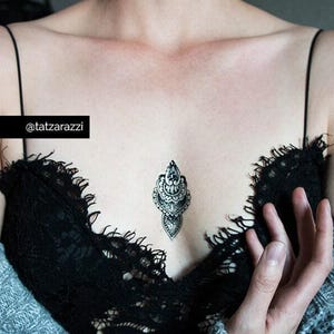 May include: A black and white temporary tattoo with an ornate design is centered on the chest. The tattoo is surrounded by a black lace top and a gray sweater. The text "@tatzarazzi" is in a black rectangle.