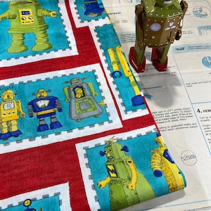 May include: A patterned fabric with cartoon robots in green, blue, and yellow, set against a red and turquoise background. A vintage green robot toy stands nearby. The fabric is likely for crafting or sewing.