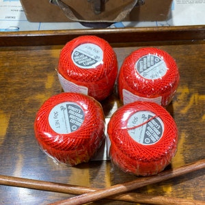May include: Four balls of vibrant red Strutt's Milford Knitting Cotton, each 50g NET. The yarn is wrapped in clear plastic. Two wooden knitting needles are present, alongside a vintage brown case.