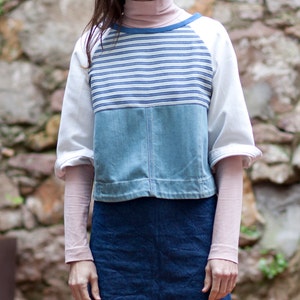 Op de afbeelding: Een wit, blauw en denim patchwork top met lange mouwen en een ronde hals. De top heeft een gestreept patroon op het bovenste deel en een denim paneel op het onderste deel. De mouwen zijn lang en hebben een witte manchet. De top wordt gedragen over een denim rok.