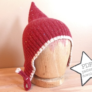 May include: A red knitted hat with a pointed crown and ear flaps. The hat features a white trim and a small decorative detail on the ear flap. It is displayed on a wooden head form. The image includes a star-shaped graphic with the text "PDF pattern download."