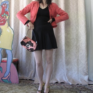 May include: A woman wearing a coral blazer, a black dress, and black tights, is holding a pink and black handbag with a gold buckle. She is standing in front of a white curtain.