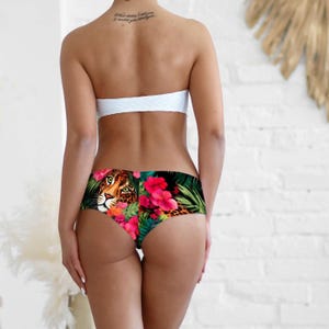 May include: A white strapless bikini top paired with bikini bottoms featuring a vibrant tropical print. The bottoms display a detailed leopard face surrounded by bright pink hibiscus flowers and lush green palm leaves against a dark background.