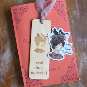 Op de afbeelding: Een houten bladwijzer met een lasergeëtste illustratie van een vrouw met vogels in haar haar en de woorden "real duck material". Een sticker met dezelfde afbeelding en tekst staat op een rood boek met bloemmotieven en een roze lint.