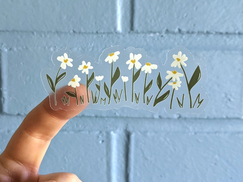 Op de afbeelding: Een transparante sticker met een ontwerp van witte madeliefjes. De sticker toont meerdere madeliefjes met gele centra en groene stelen en bladeren.