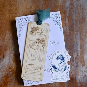 May include: A rectangular wooden bookmark featuring a laser-engraved illustration of a bookshelf with a sleeping cat, potted plants, and stacks of books, topped with a teal fabric ribbon, resting on a white book with a blue sticker reading Dangerous Reader.
